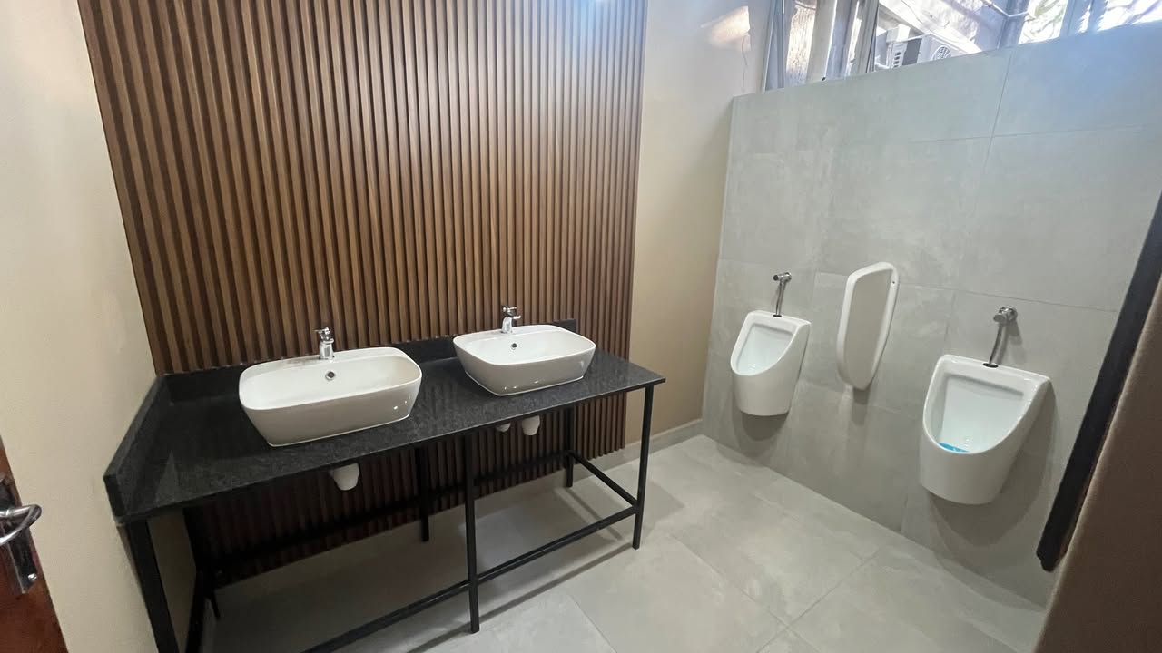 Modern commercial men's restroom with double vessel basins on granite-top vanity, slatted wood feature wall and wall-mounted urinals by Plumbing Beat