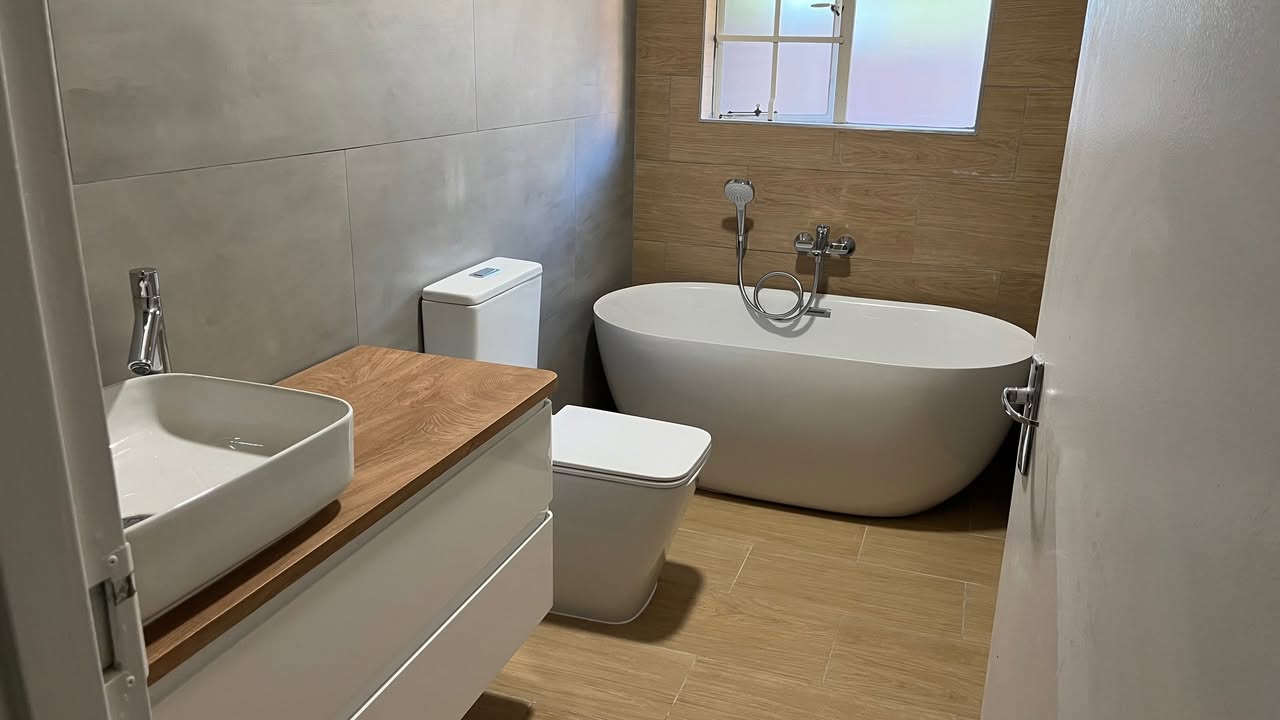 Modern compact bathroom with freestanding oval tub, wood-look floor tiles, wall-hung vanity and vessel basin by Plumbing Beat