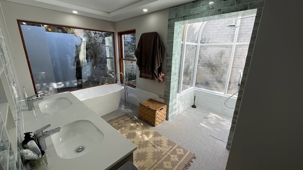 Spacious spa-style bathroom renovation by Plumbing Beat featuring a freestanding bathtub, double vanity basin, green-tiled walk-in shower with rainfall showerhead, and large garden-facing windows